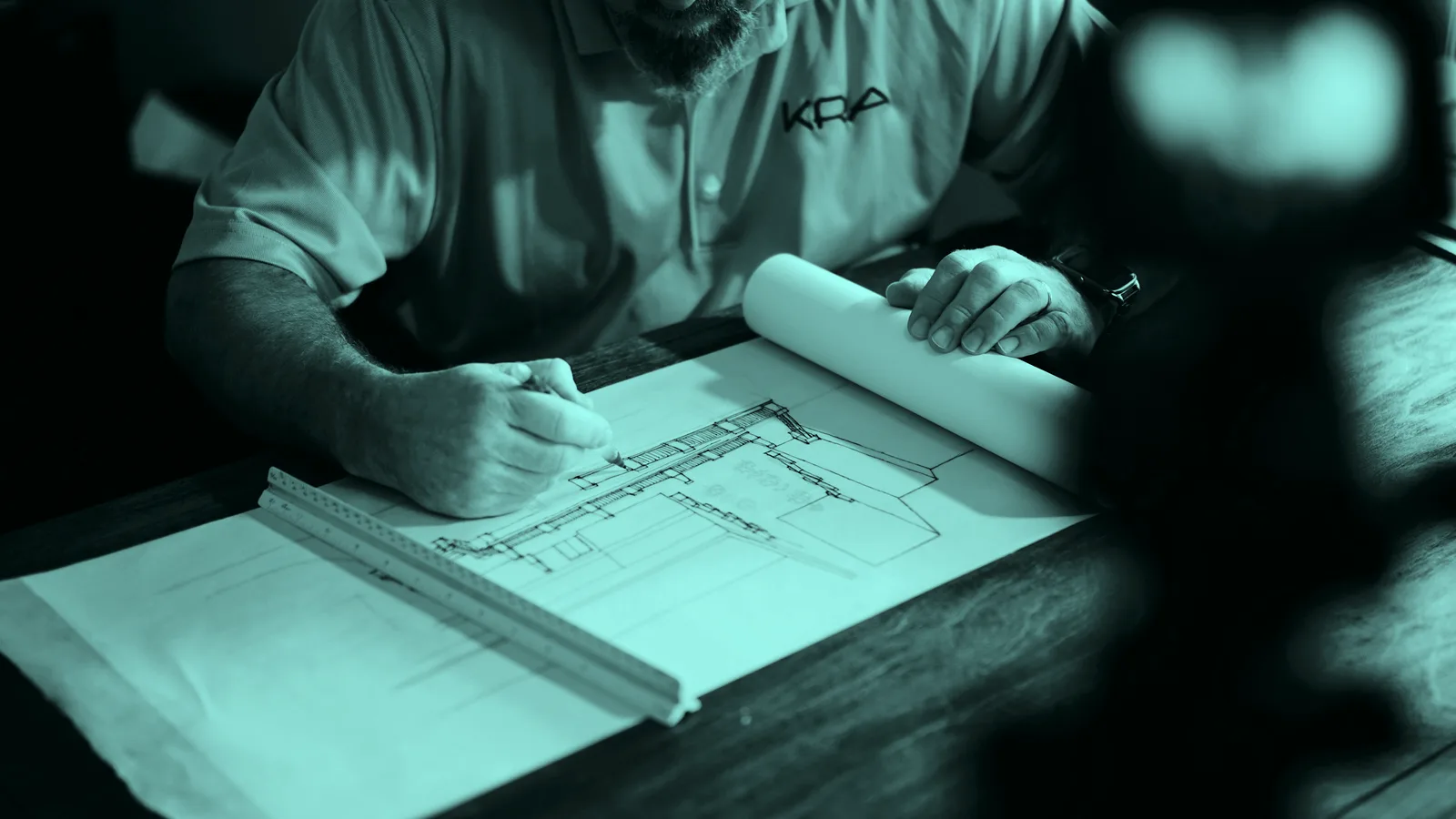A hand drafting on plans with a pencil and ruler, tinted teal